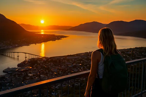 A blonde woman with a backpack stands on a viewing terrace above Tromsø, looking at the city and the Tromsøysund Bridge in the golden light of the midnight sun; in the background, calm water, mountains, and the illuminated buildings of the city